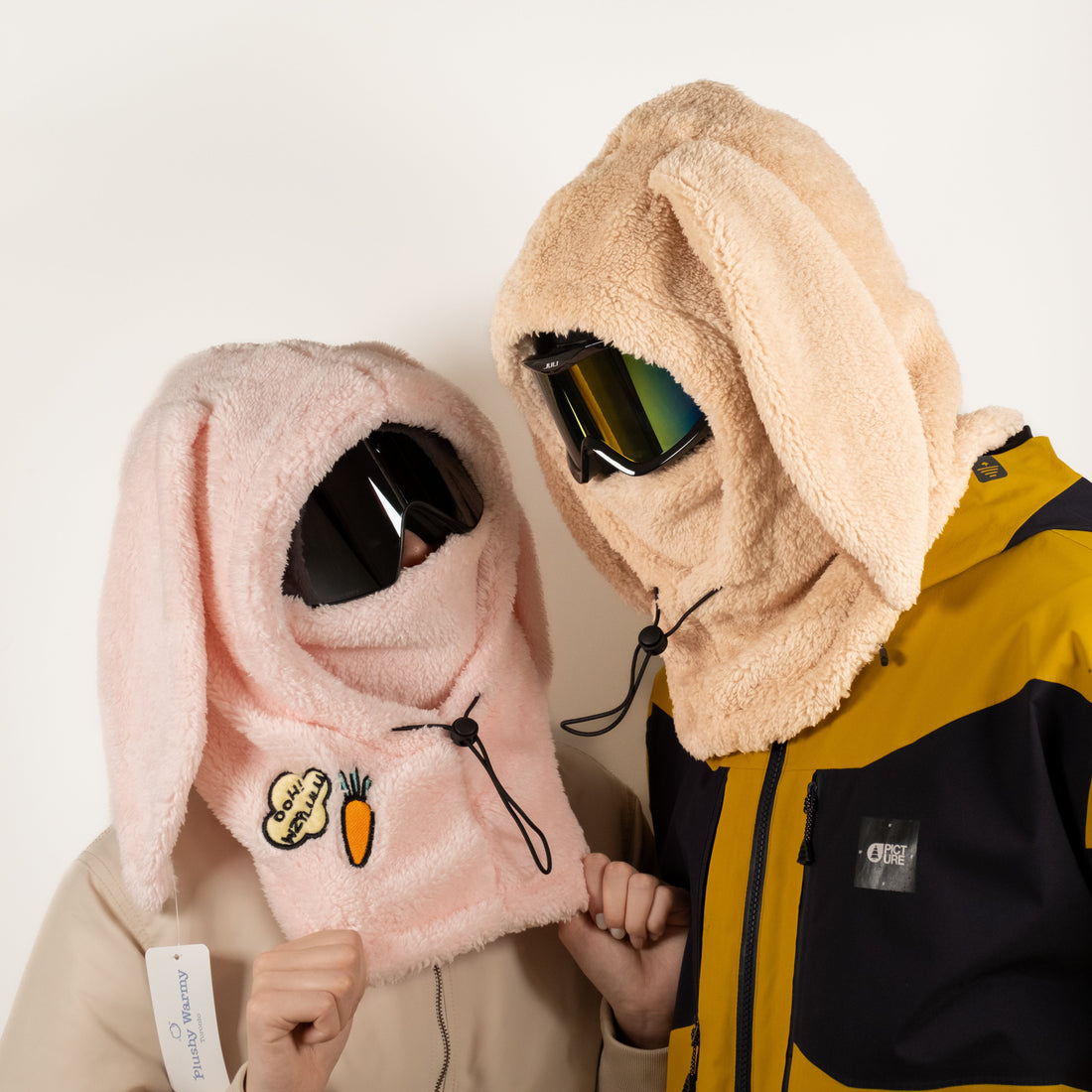Two skiers wearing pink and beige bunny plush helmet hoods with ski goggles, showing the adjustable drawcord toggle